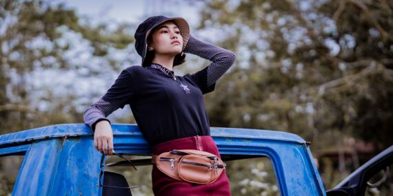 Woman in a black bucket hat, layered black top, burgundy skirt, and waist bag posing beside a blue jeep.
